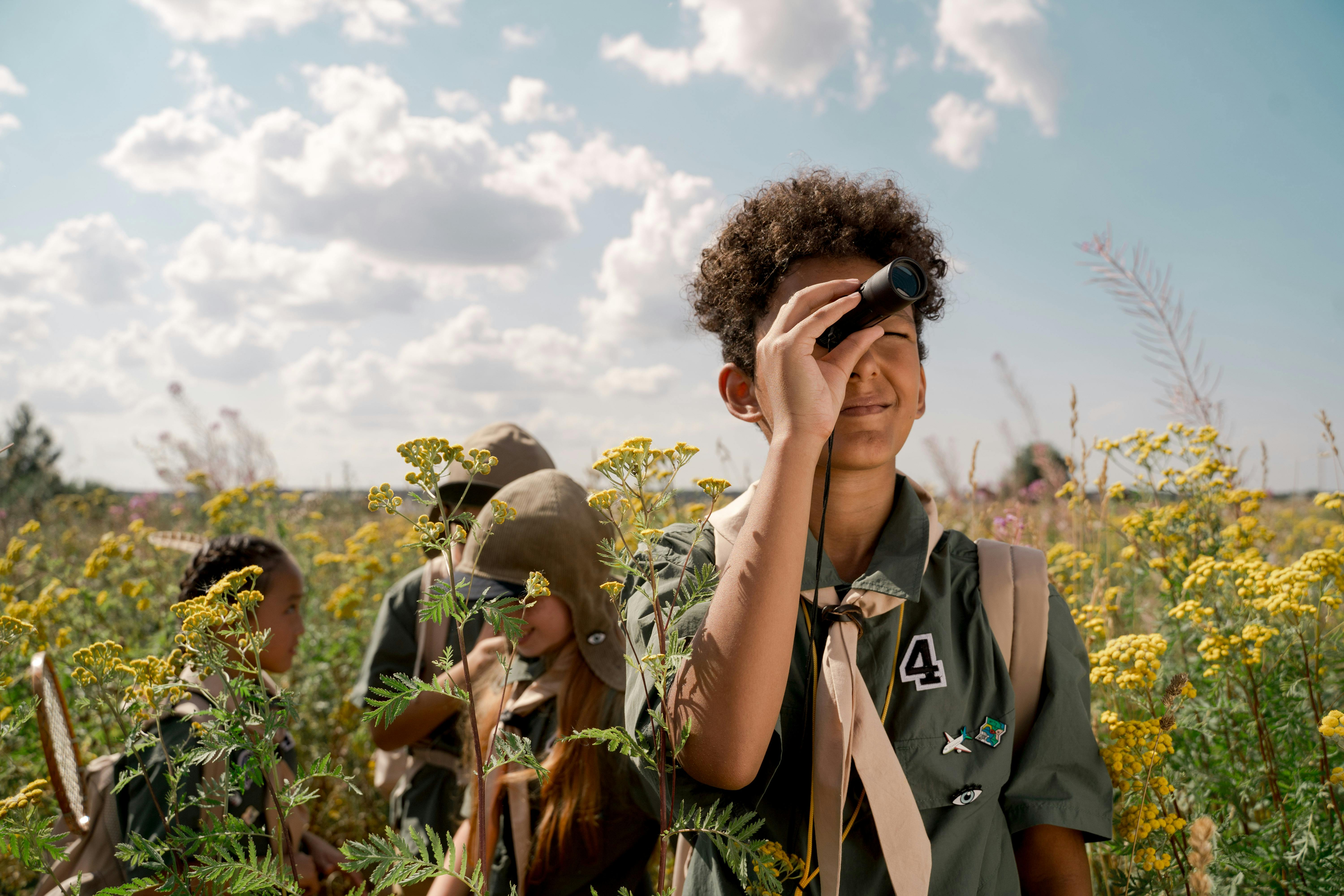 Children in scout uniforms exploring a field with binoculars and backpacks